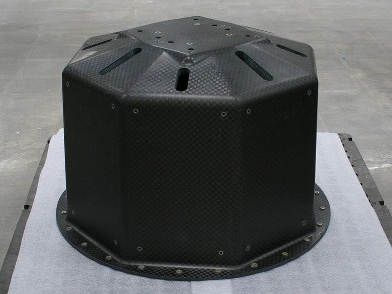 Image shows the octagonal section of the smallsat launch adapter structure in a manufacturing area and looks down at it from slightly above and to the side. It is sitting on a dark-colored pallette with a protective white foam layer on its surface. The surface is a matte carbon fiber weave. There are metallic inserts, cutouts and drill holes at various junctures. 